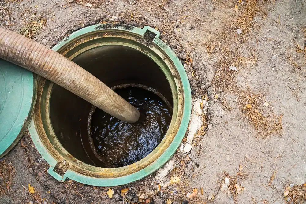 A large hose is inserted into an open green septic tank, pumping out wastewater. The surrounding ground is dry with some leaves and dirt scattered around the tank.