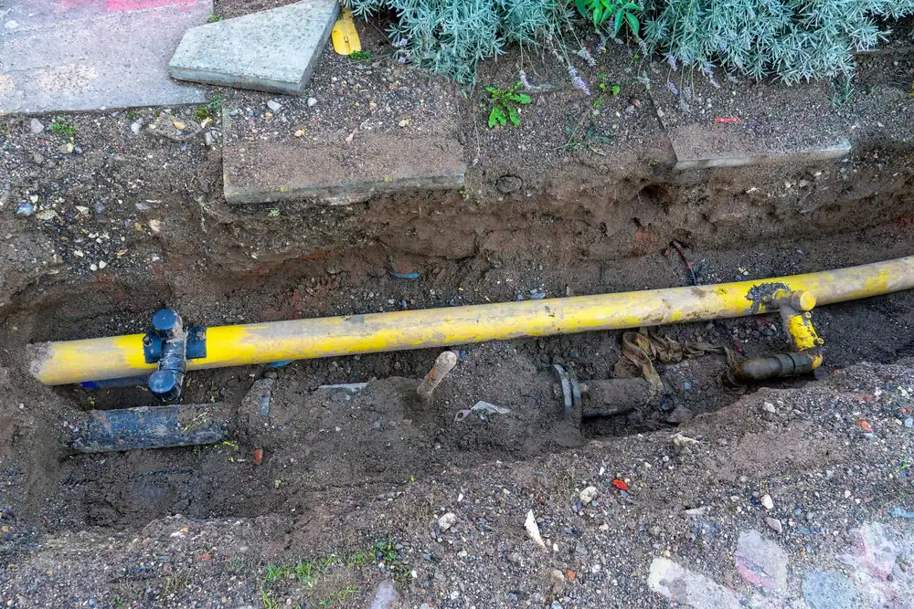 A yellow gas pipeline is exposed in a dug trench surrounded by soil and gravel, with pipes and fittings visible. Stone slabs and green plants are at the edge of the excavation.