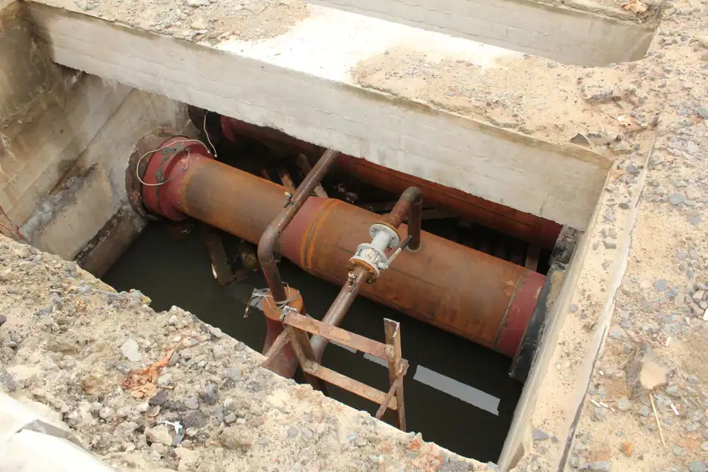 Large, rusted industrial pipes are exposed in a rectangular pit filled with water, surrounded by concrete and sandy soil, possibly part of construction or repair work.