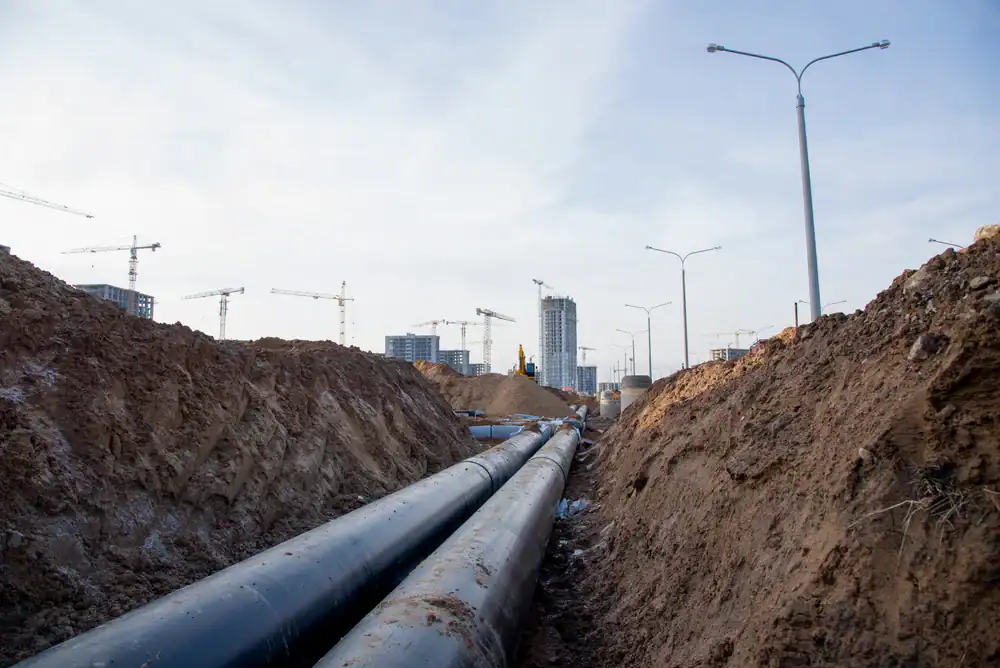 Large black pipes are laid in a trench at a construction site, with dirt mounds on each side. City buildings and numerous cranes are visible in the background under a cloudy sky.